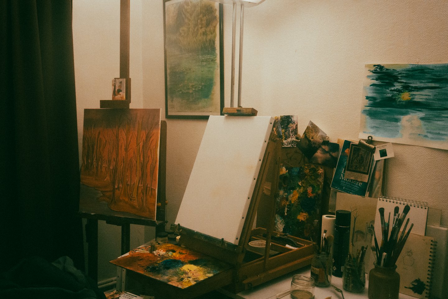 Art studio with paintings, brushes, and palette on desk.
